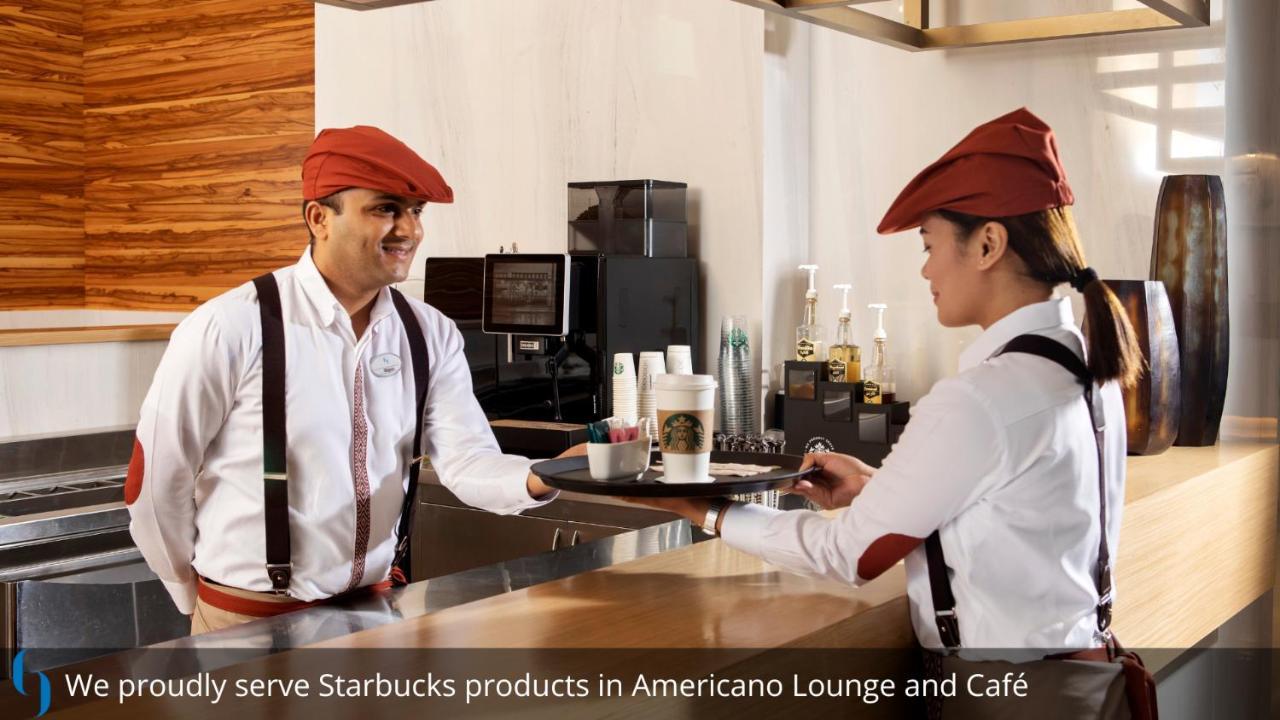 Grand Cosmopolitan Hotel Dubai Exteriör bild The photo shows two staff members in a café environment. One is handing a tray with a Starbucks beverage to the other, who is standing at the counter. Both employees are wearing white shirts with suspenders and red caps. The background suggests a mod